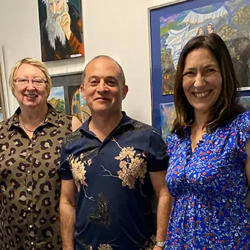 Zwei Frauen und ein Mann stehen vor einer Bilderwand und lächeln