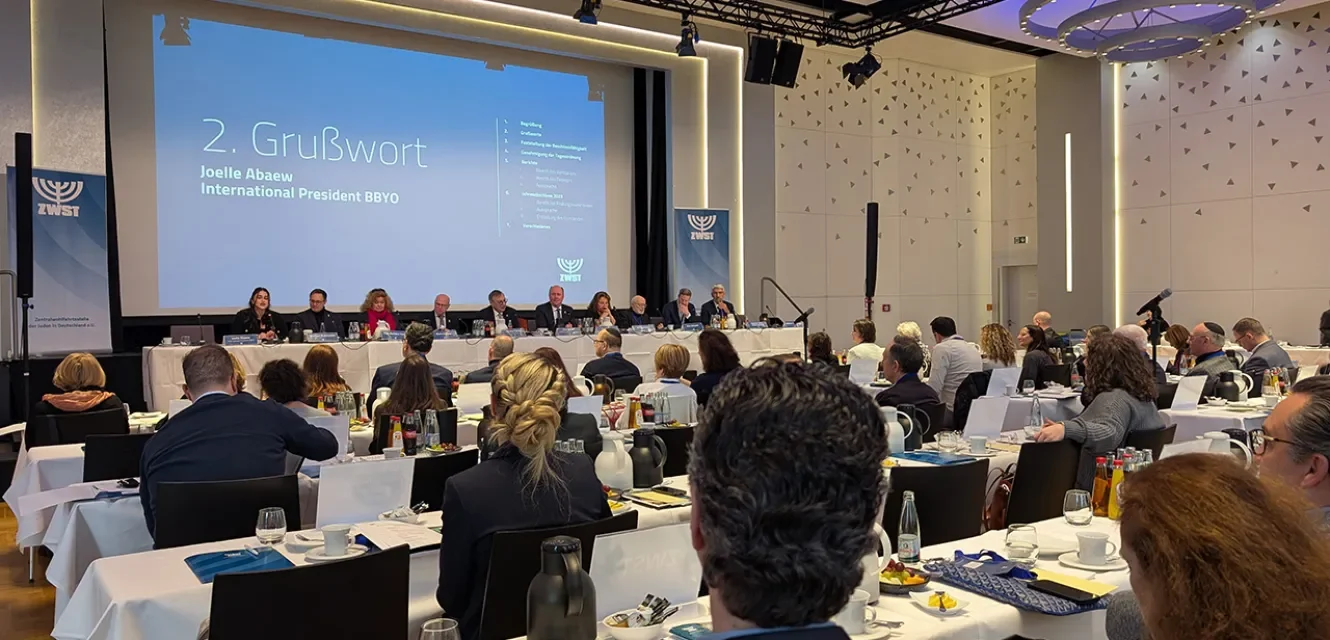 Plenum in einem Saal voller Tischreihen mit Publikum